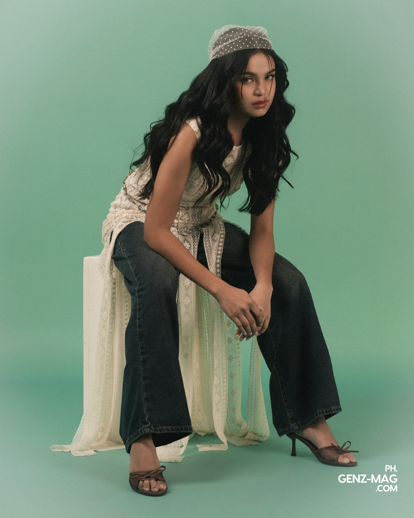 Alt text: "Zephanie perched on a white pedestal in a lace top with fringe detailing and wide-leg denim, wearing brown strappy heels, for GEN Z Magazine's cover story with Michael Sager