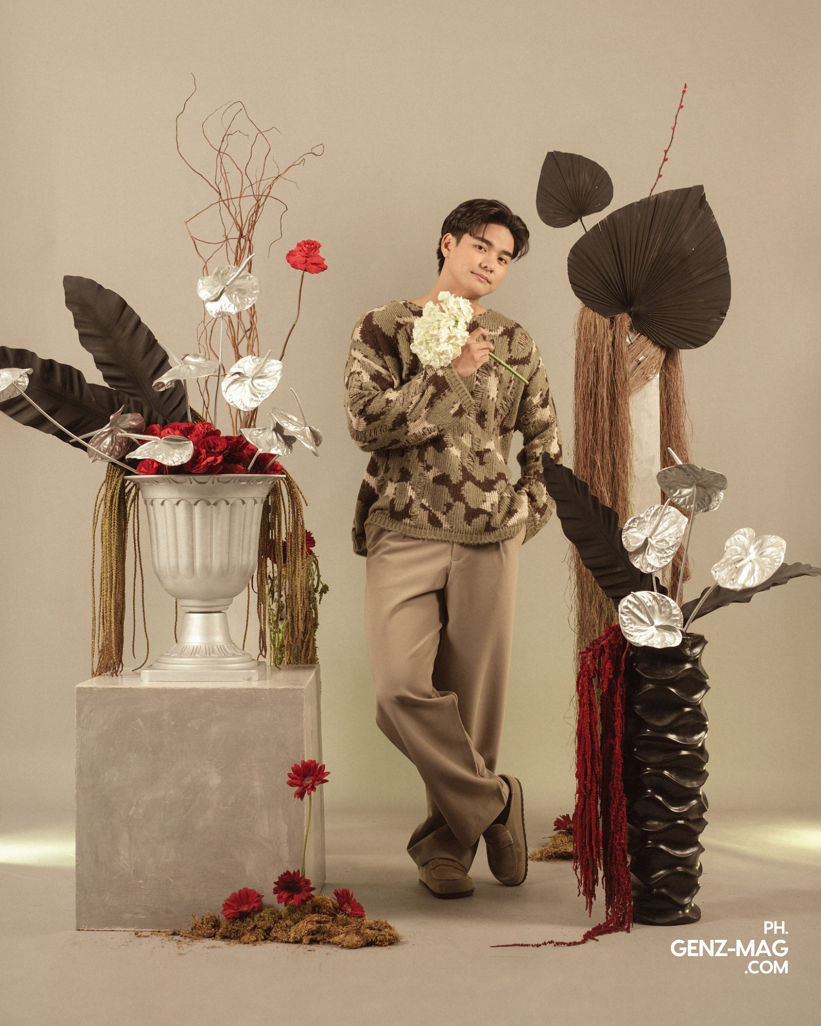 Michael Sager in a knit sweater holding a white flower, standing among a dried floral and foliage set by Makati Flower Atelier, for GEN Z Magazine's cover story with Zephanie